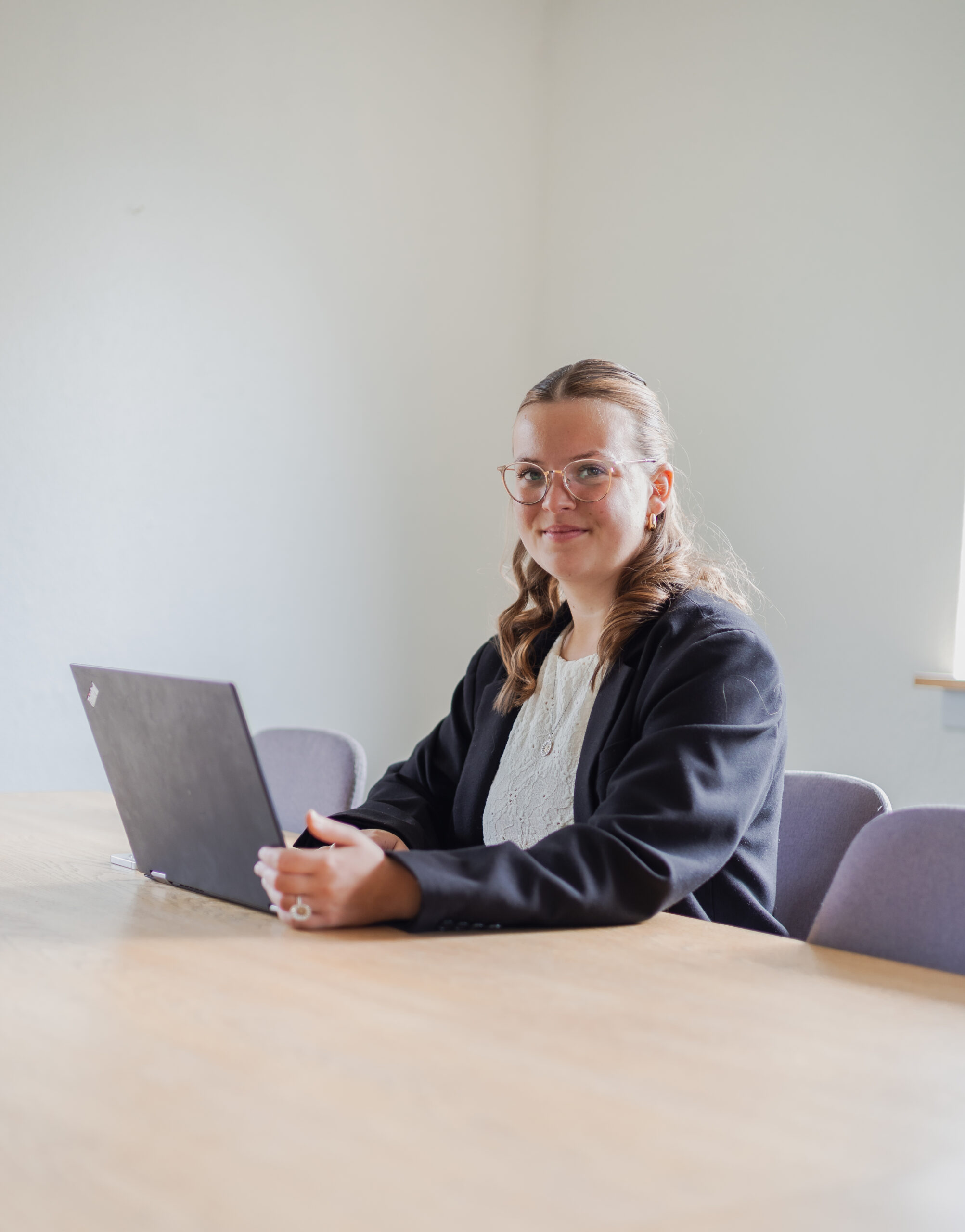Professionel kvinde i sort blazer sidder ved mødebord med bærbar computer.