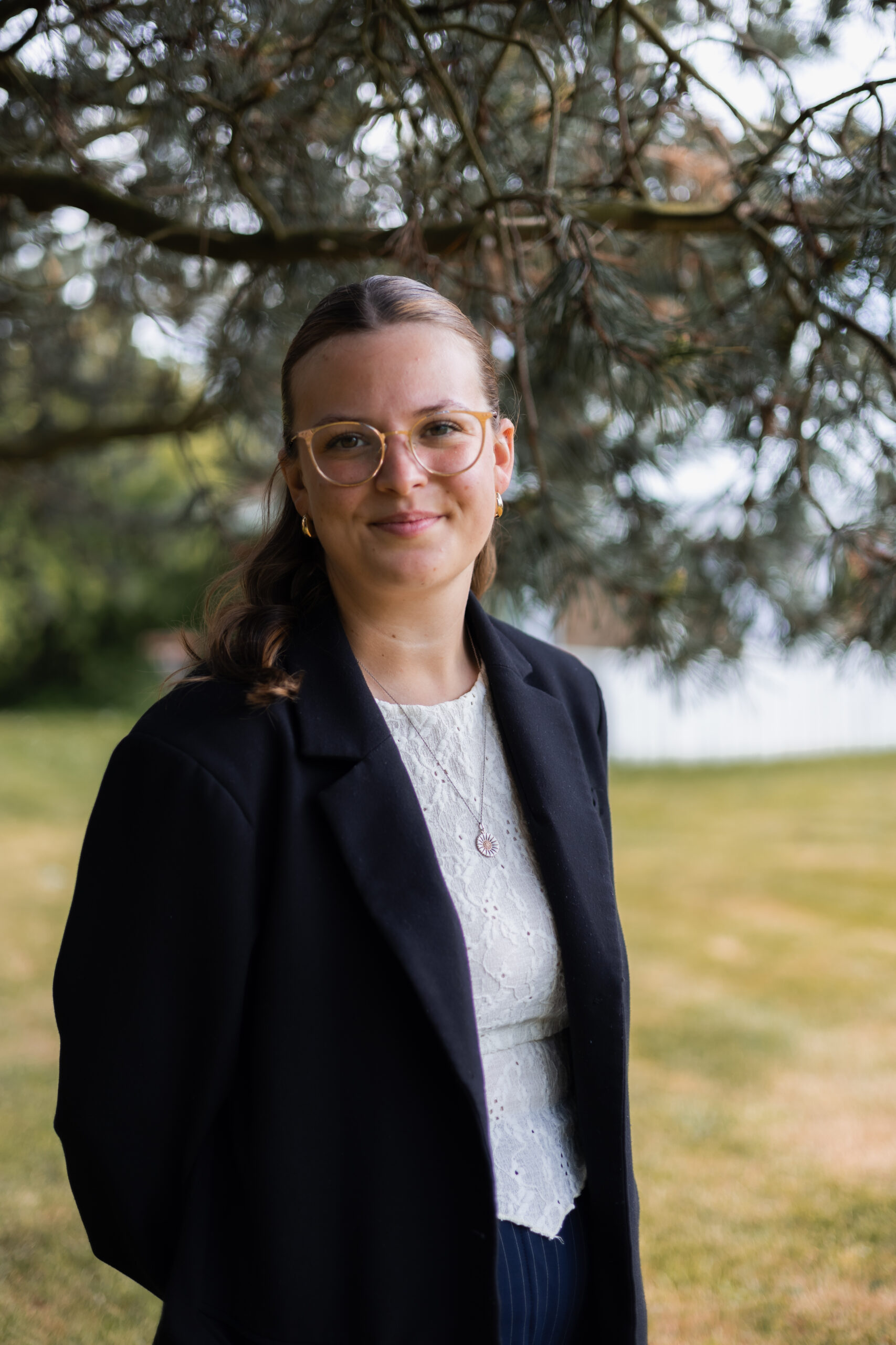 Portræt af professionel kvinde i sort blazer og hvid blondebluse, stående udendørs under et træ.